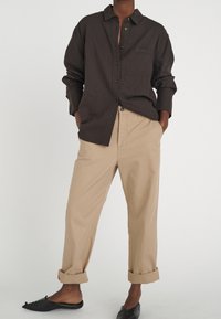 Brown button-up shirt with a chest pocket, paired with beige cuffed pants and black slip-on shoes. Smooth fabric textures visible.
