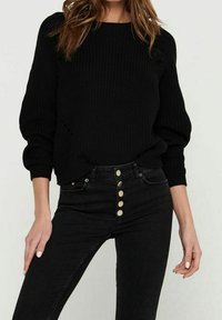 Schwarzer Strickpullover mit rundem Halsausschnitt und Ballonärmeln, kombiniert mit hochgeschnittenen schwarzen Jeans, die eine Reihe von fünf goldenen Knöpfen aufweisen.