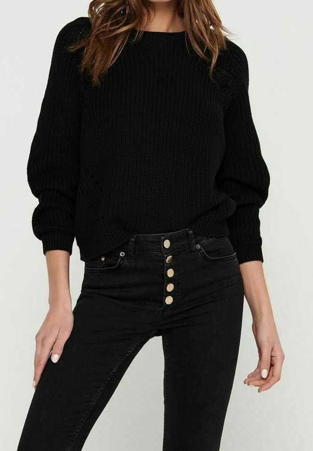 Schwarzer Strickpullover mit rundem Halsausschnitt und Ballonärmeln, kombiniert mit hochgeschnittenen schwarzen Jeans, die eine Reihe von fünf goldenen Knöpfen aufweisen.