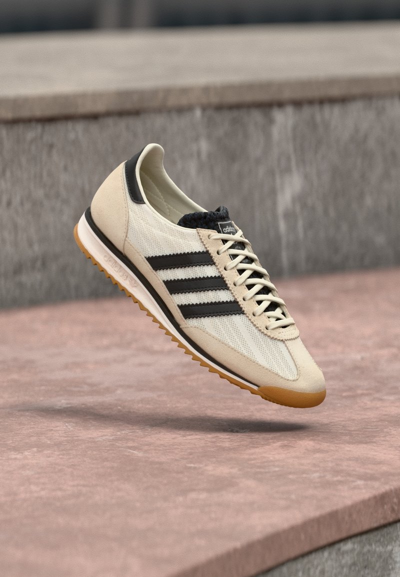 Μπεζ και μαύρο αθλητικό παπούτσι Adidas με σόλα από καουτσούκ, αιωρείται πάνω από μια πέτρινη επιφάνεια, δείχνοντας πλευρικές και επάνω όψεις.