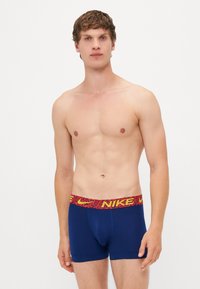 Mørkeblå boxershorts med en rød og gul mønstret talje med "NIKE" logo. Blødt stof, tætsiddende design og længde til midten af låret.