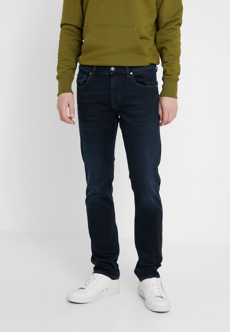 Mörkblå denimjeans med smal passform, fem fickor, subtila sömnadsdetaljer och kompletterat med en grön hoodie och vita sneakers.