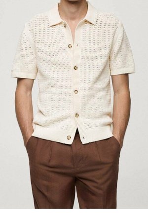 Homme portant une chemise à manches courtes en tricot crème avec col et boutons, et un pantalon brun, mains dans les poches, sur un fond uni.