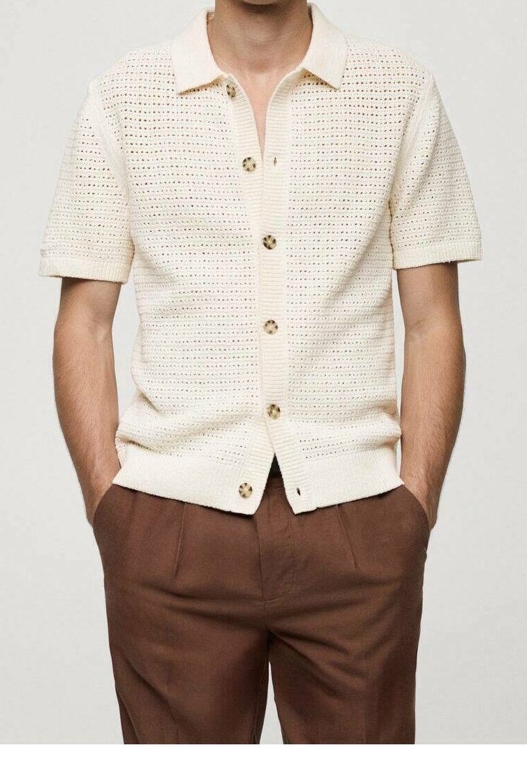 Homme portant une chemise à manches courtes en tricot crème avec col et boutons, et un pantalon brun, mains dans les poches, sur un fond uni.