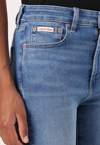 Lichtblauwe denim jeans met een hoge taille, voorzien van een rood-witte Calvin Klein logo-label, metalen knopen en een soepele textuur.