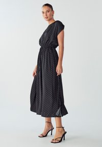 Black polka dot dress with short sleeves, gathered waist, and flowing skirt. Accompanied by black strappy heeled sandals.