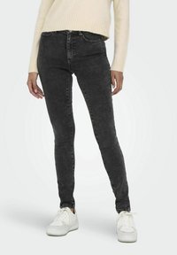 Svarta skinny jeans i denim, med hög midja och subtila slitningar, i kombination med vita sneakers och en krämfärgad tröja.