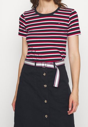 Vrouw draagt een donkerblauw, wit en rood horizontal gestreept T-shirt, ingestopt in een zwarte, hooggetailleerde rok met knopen en een witte riem.