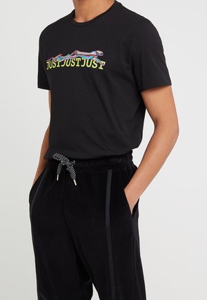 Mężczyzna w czarnej koszulce z kolorowym biegnącym gepardem nad powtarzającym się żółtym napisem "JUST" oraz w czarnych spodniach z troczkami, z rękami w kieszeniach.