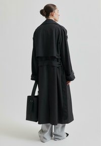 Długi czarny płaszcz w stylu trencza z paskiem w talii, szerokim kołnierzem i detalami w postaci klap. Zestawiony z jasnoszarymi spodniami o szerokich nogawkach oraz czarną torbą na ramię.