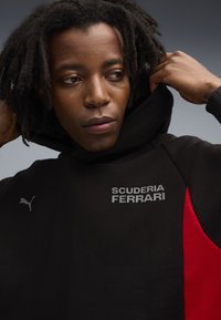 Sort hættejakke med røde detaljer, prydet med "SCUDERIA FERRARI" i sølv tekst og et Puma-logo. Lavet af tekstureret stof og designet med en hætte.