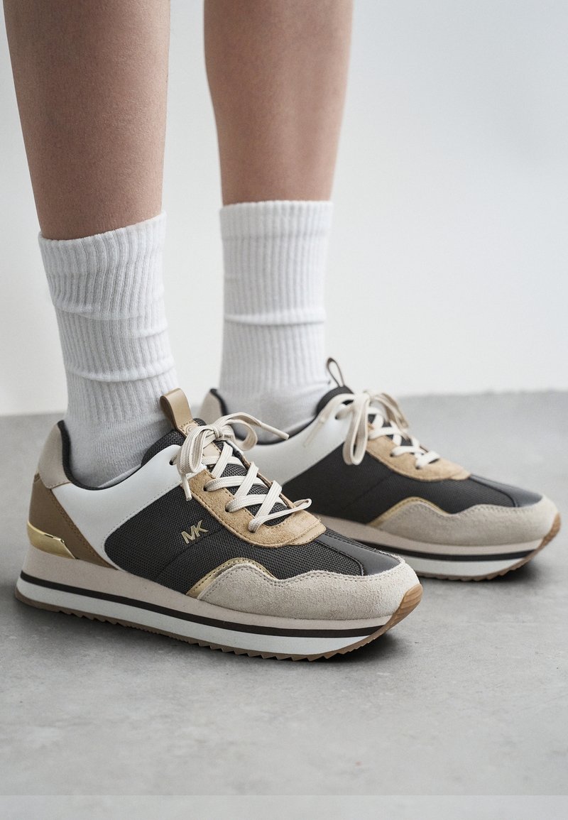 Jambes portant des chaussettes blanches et des baskets Michael Kors beiges, noires et blanches, debout sur un sol gris.