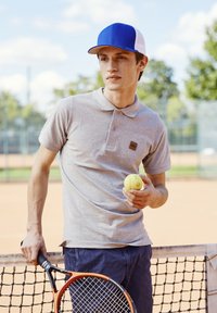 Grå pikétröja med krage och logotyp, blå och vit keps, håller en tennisboll, lutar sig mot ett tennisracket vid ett nät.