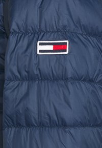 Veste matelassée bleu marine avec un patch logo rectangulaire présentant des blocs de couleur horizontaux en noir, blanc et rouge.