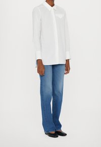 Weißes langärmliges Hemd mit Button-down-Kragen und Brusttasche, kombiniert mit blauen Jeans im geraden Schnitt und schwarzen Schuhen.