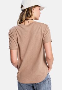 Vrouw in een beige shirt met korte mouwen, lichtblauwe jeans, een witte pet en een schelpketting, met haar rug naar de camera gekeerd en met haar handen in de zakken.
