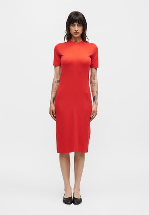 Femme aux cheveux bruns mi-longs portant une robe rouge ajustée arrivant aux genoux et des chaussures plates noires, debout devant un fond clair uni.