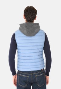 Uomo che indossa un gilet trapuntato blu chiaro con cappuccio grigio, una maglietta a maniche lunghe blu navy e jeans blu scuro, visto da dietro.