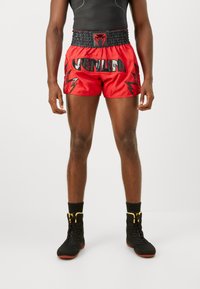 Red satin boxing shorts with black elastic waistband, featuring bold black text and lightning bolt accents. Paired with black athletic boots.