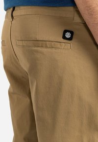 Khaki shorts gemaakt van een katoenen mengsel, met een vlakke voorkant, achterzakken en een zwart logo-opdruk op de rechter achterkant.
