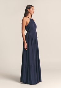 Lång mörkblå klänning i lätt tyg med halterneck, figursydd midja och flerskiktad, utsvängd kjol med volangdetaljer.