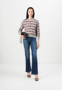 Grauer Pullover mit rotem und schwarzem Logo-Muster, kombiniert mit hochgeschnittenen blauen Schlagjeans und schwarzen Riemchensandalen, während sie eine schwarze Tasche hält.