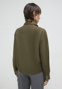 Olivegrüne Bluse mit langen Ärmeln, glatter Textur, Kragen und Knopfmanschetten. Kombiniert mit grauen Nadelstreifen-Hosens.