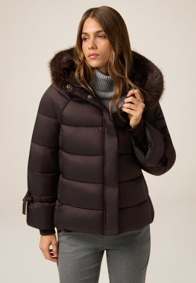 Chaqueta de plumas marrón con capucha desmontable forrada de piel, costuras horizontales y puños ajustables. Combinada con un suéter de cuello alto gris.