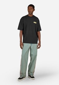 Melnas kokvilnas t-kreklas ar apaļu kakla izgriezumu un mazu zelta logotipa detaļu uz krūtīm, savienots ar gaiši ziliem džinsu biksēm ar sānu svītrām.