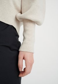 Svetlosmeđi pleteni pulover s opuštenim krojem, s rebrastim manžetama. Ispod je crna tkanina, izbliza prikazana ruka koja se odmara na šavu.