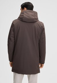 Mann in einem dunkelbraunen Wintermantel mit Kapuze, von hinten gesehen vor einem schlichten weißen Hintergrund.