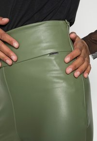 Falda verde de imitación cuero de cintura alta con textura suave, que presenta un detalle de costura y un pequeño acento de logo plateado en la cinturilla.