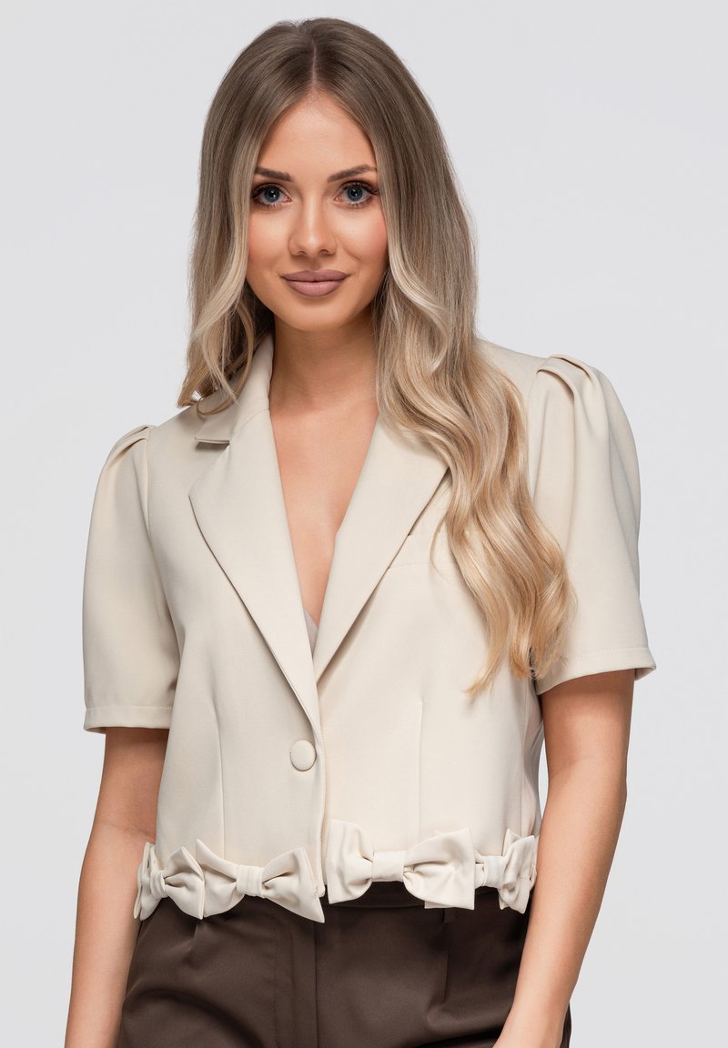 Jeune femme aux longs cheveux blonds ondulés porte un blazer crème à manches courtes avec des détails en nœud et un pantalon sombre, debout sur un fond uni.