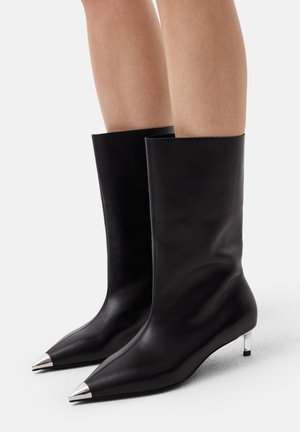 Black mid-calf leather boots with pointed silver toe caps and slim silver kitten heels worn on bare legs against a white background.