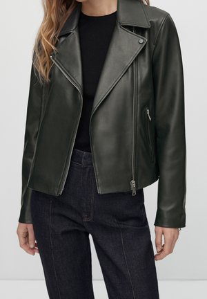 Femme porte une veste en cuir de motard vert foncé sur un haut noir avec un jean taille haute foncé sur un fond uni.