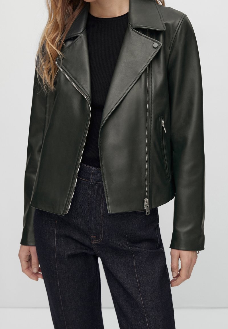 Femme porte une veste en cuir de motard vert foncé sur un haut noir avec un jean taille haute foncé sur un fond uni.