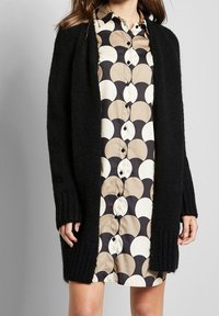 Czarny dziergany kardigan z żebrowanymi mankietami, z otwartym przodem, noszony na beżowo-czarnej sukience koszulowej z wzorem w kółka.