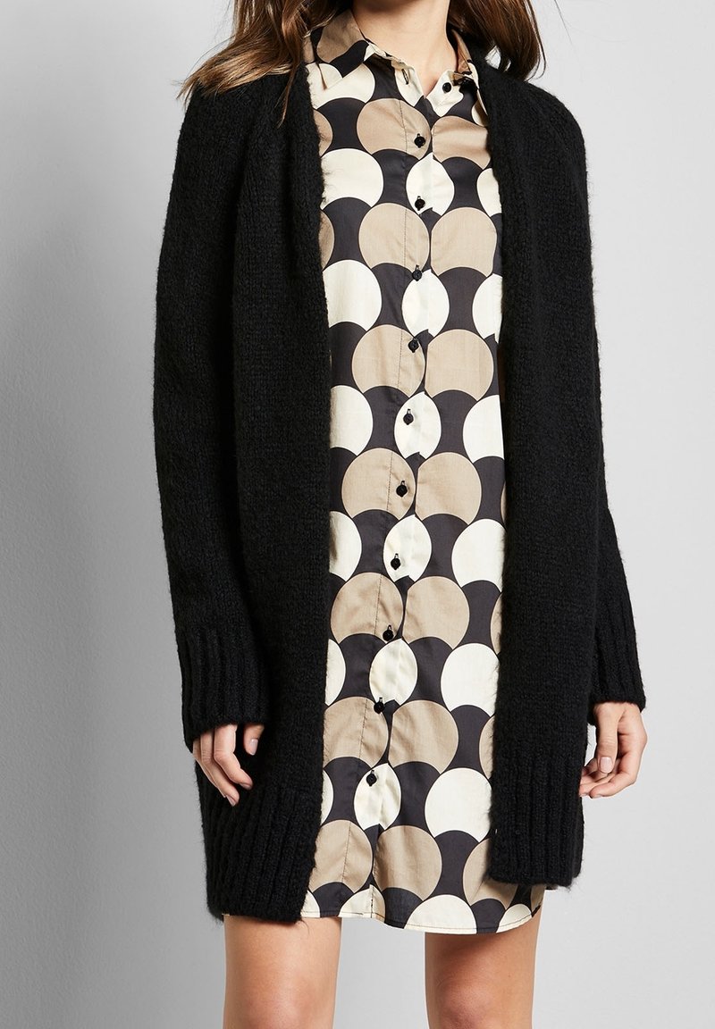 Czarny dziergany kardigan z żebrowanymi mankietami, z otwartym przodem, noszony na beżowo-czarnej sukience koszulowej z wzorem w kółka.