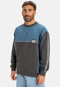 Felpa girocollo con blocchi di colore blu e grigio, un logo sul fronte e strisce laterali a fantasia. Tessuto morbido con vestibilità rilassata.