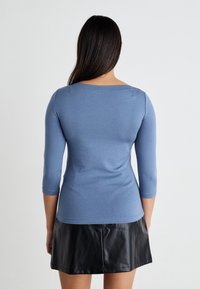 Μπλε, εφαρμοστό τοπ με μακρύ μανίκι και τετράγωνη λαιμόκοψη, φτιαγμένο από μαλακό, ελαστικό ύφασμα. Συνδυασμένο με μια μαύρη δερμάτινη μίνι φούστα.