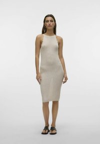 Abito lavorato a maglia beige con una silhouette aderente, scollatura a forma di scallop e lunghezza a metà polpaccio. Indossato con sandali neri flat, su uno sfondo semplice.