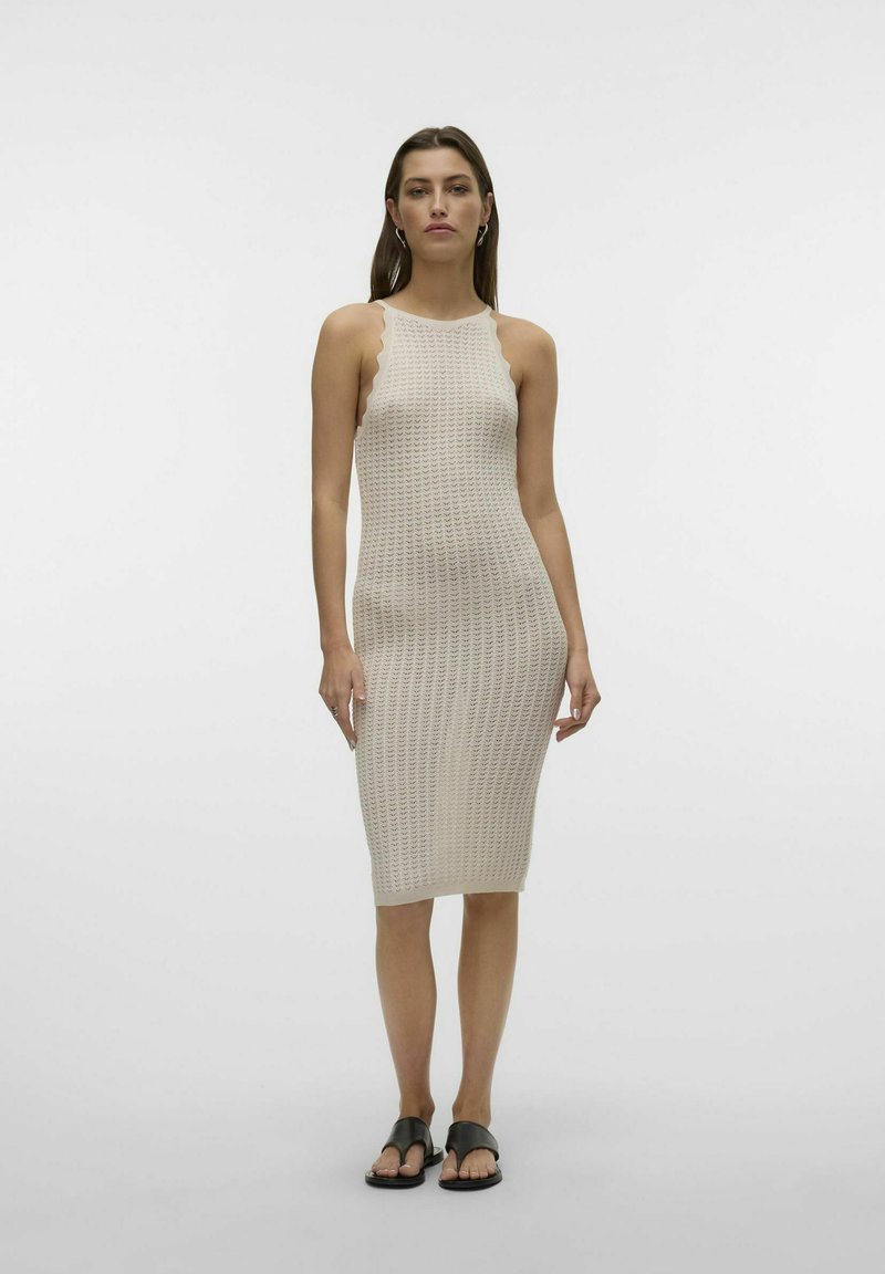 Abito lavorato a maglia beige con una silhouette aderente, scollatura a forma di scallop e lunghezza a metà polpaccio. Indossato con sandali neri flat, su uno sfondo semplice.