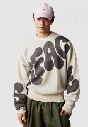 Jeune homme portant un pull beige avec de grandes lettres abstraites foncées et une casquette rose clair, debout avec une main dans la poche d’un pantalon vert.
