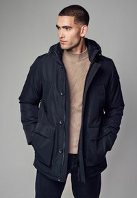 Mustapukuinen parka-takki huppuineen, jossa on suuret etutaskut, vetoketjullinen etuosa ja sileä pinta. Puettu beige-kuiten neuleen päälle.