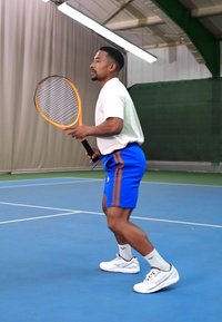 Tennisimängija valges särgis ja sinistes lühikestes pükstes, millel on oranžid triibud, hoiab käes reketit. Tal on jalas valged tossud sinisel väljakul.