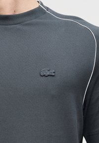 T-shirt de sport gris foncé en tissu texturé, avec un petit logo embossé sur le côté gauche de la poitrine et des passepoils blancs le long des coutures.
