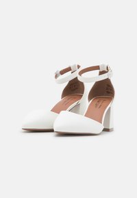 Chaussures blanches à talons hauts avec un bout pointu, une bride au niveau de la cheville et un talon carré. Matériau lisse avec une semelle intérieure beige et des détails de couture subtils.