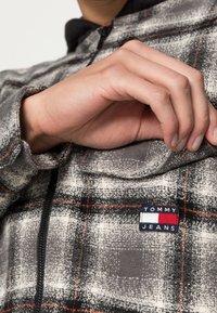 Grijze en zwarte geruite jas van gestructureerde stof, met een ritssluiting aan de voorkant en een gestikt logopatch in rood, wit en blauw op de zak.