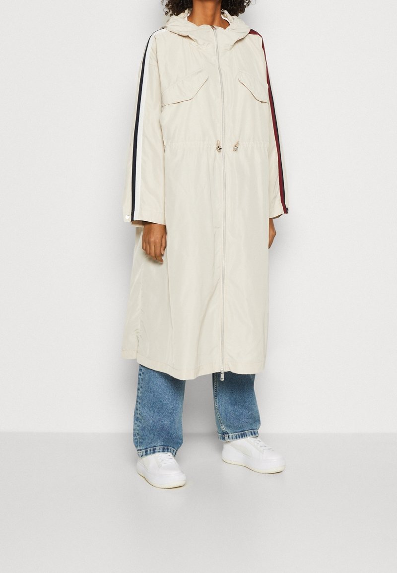 Mujer con un largo abrigo con capucha color crema con rayas negras y rojas en las mangas, vaqueros azules y zapatillas blancas, de pie sobre un suelo gris.