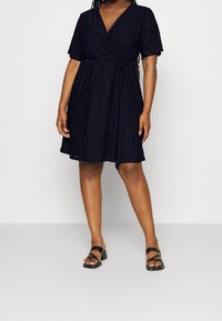 Nainen, jolla on päällään lyhythihainen tummansininen kietaisumekko, jossa on vyötärökiristys, ja mustat korkokengät, seisoo yksinkertaista taustaa vasten.